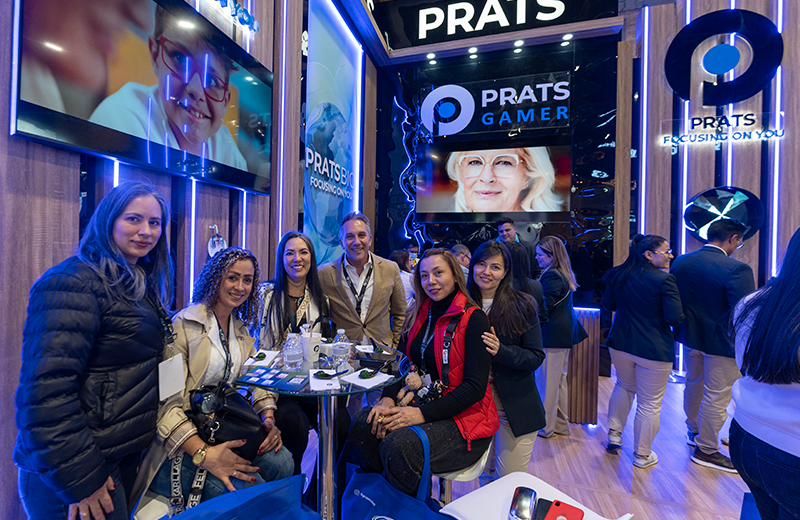 Group of people posing around a round table at a tech expo booth with blue lighting and 'PRATS GAMER' branding in the background.