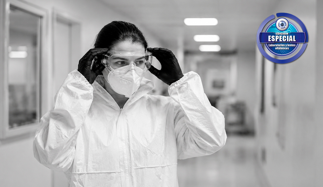 Female researcher in a white protective suit and gloves adjusts safety goggles in a sterile lab hallway, logo badge visible in the top-right corner, neutral grayscale.