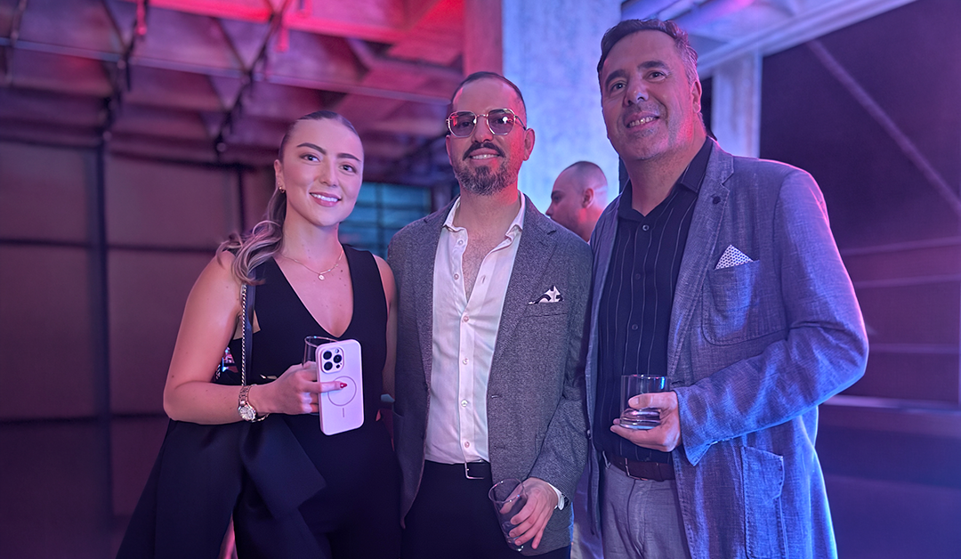 Three well-dressed people pose for a photo at a social event, each holding a drink.
