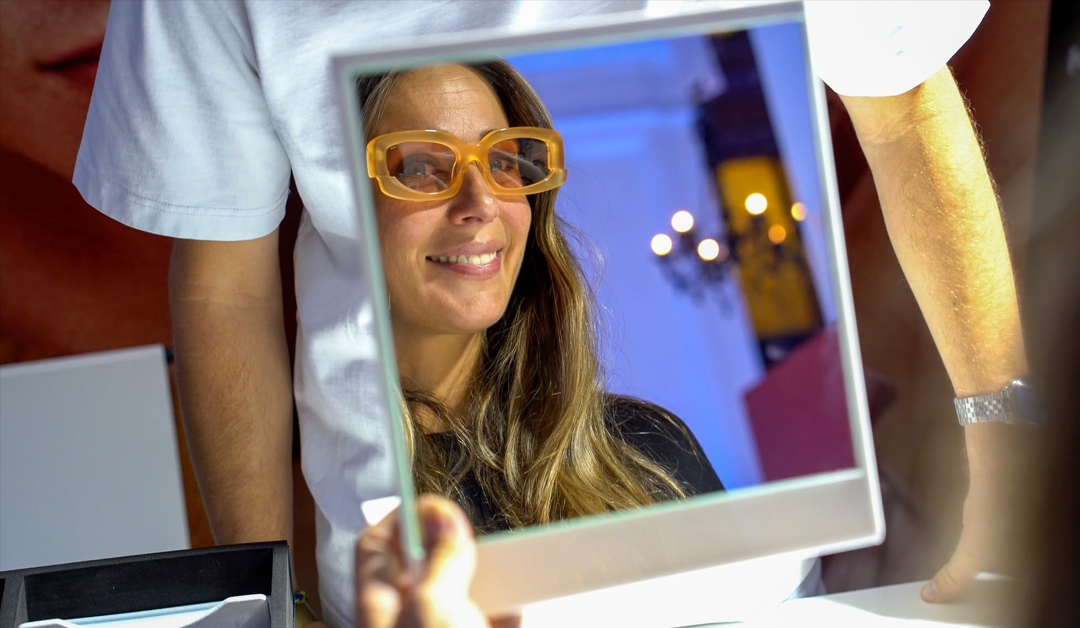 Smiling woman wearing orange safety goggles looks through a square mirror frame held in front of her.