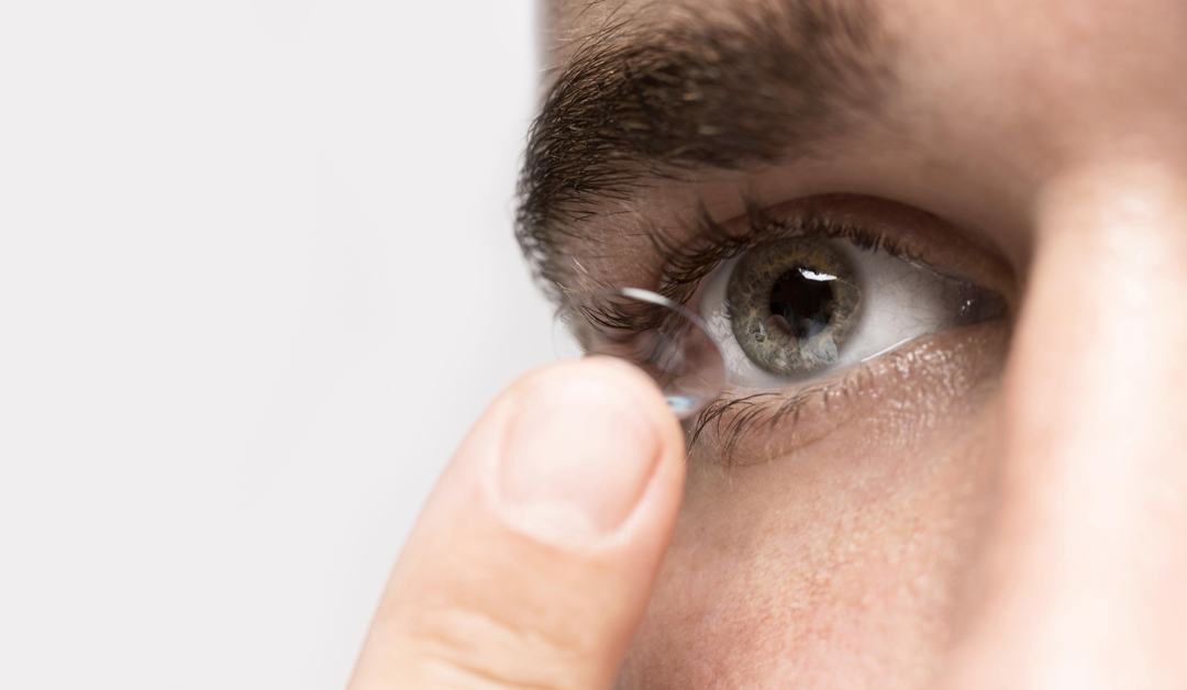 Close-up of a person inserting a contact lens into their eye, fingers holding a clear lens near the pupil.