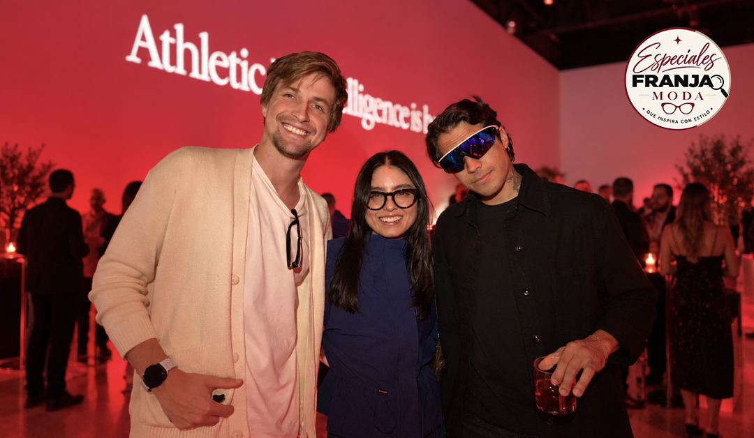 Three people posing for a photo at a fashion event with red lighting and brand logos visible in the background (Athletic... wall and a circular logo).