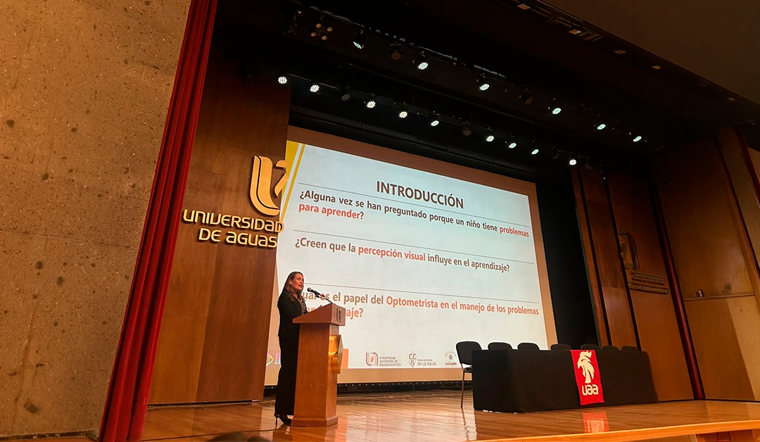 Speaker at a podium on a stage delivering a presentation; a large projection slide in Spanish is shown, with a red curtain and panel table nearby in a conference setting.