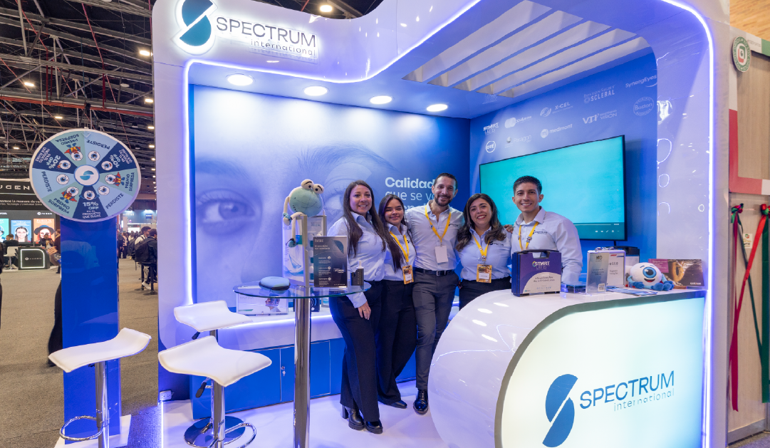 Exhibit booth for Spectrum International with a blue, curved reception desk and a group of five staff posing for photos near a large screen backdrop.