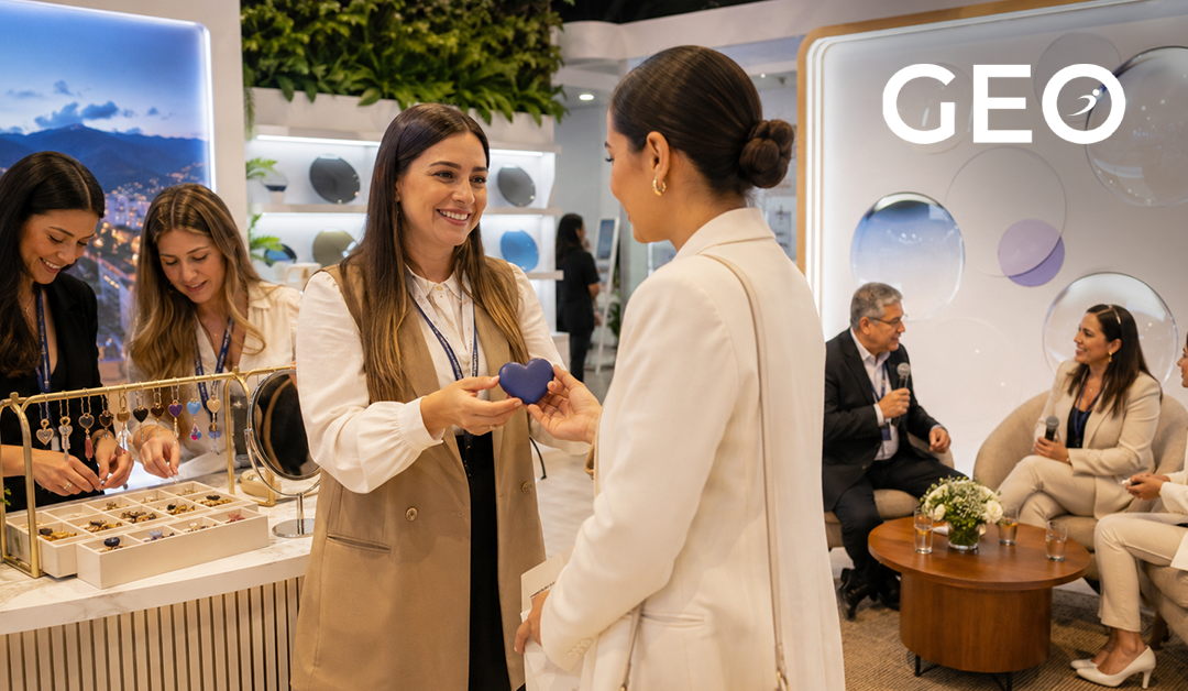 Two women at a booth exchange a blue heart object while jewelry displays glint nearby; GEO logo visible in the background.