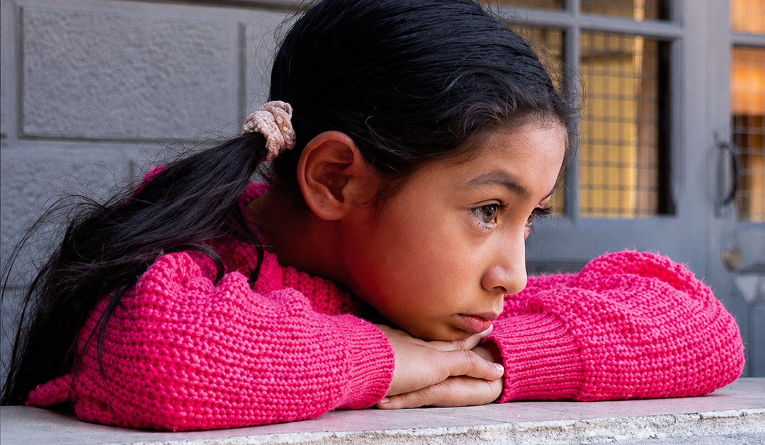 Young girl with long dark hair in a bright pink knit sweater rests her chin on folded arms on a windowsill outside a building window behind her.