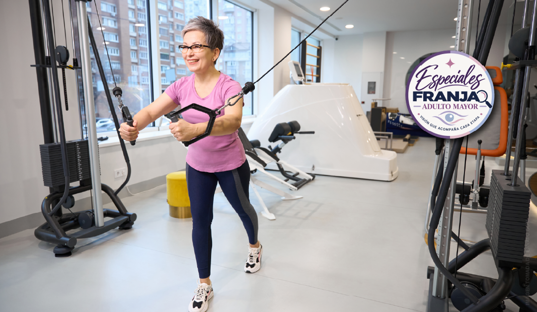 Senior woman in a pink shirt uses a cable machine in a bright gym, smiling during the exercise.