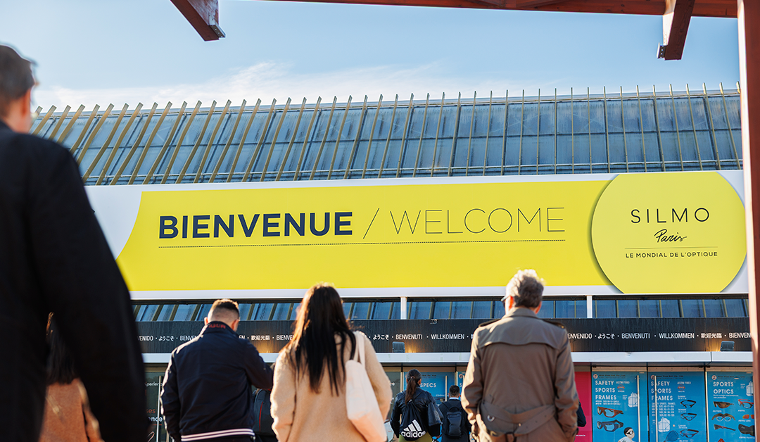 Entrance to a venue beneath a bright yellow 'Bienvenue/Welcome' sign as people walk inside