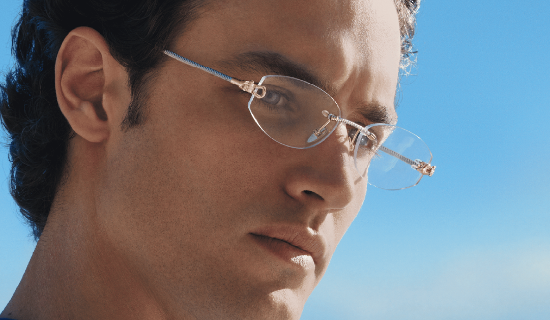 Close-up portrait of a person wearing thin metal-framed glasses outdoors with a bright blue sky behind.